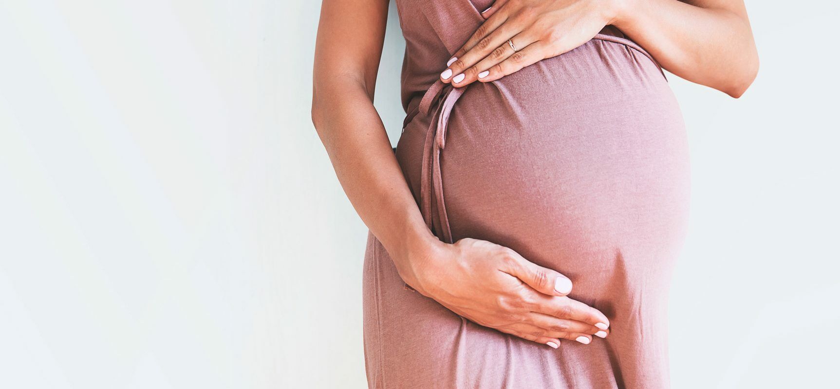 Cesarskie cięcie czy poród naturalny? Jakie są zalety porodu siłami natury i jak zadbać o noworodki urodzone przez CC? Kobieta w zaawansowanej ciąży w różowej sukience dotyka brzucha.