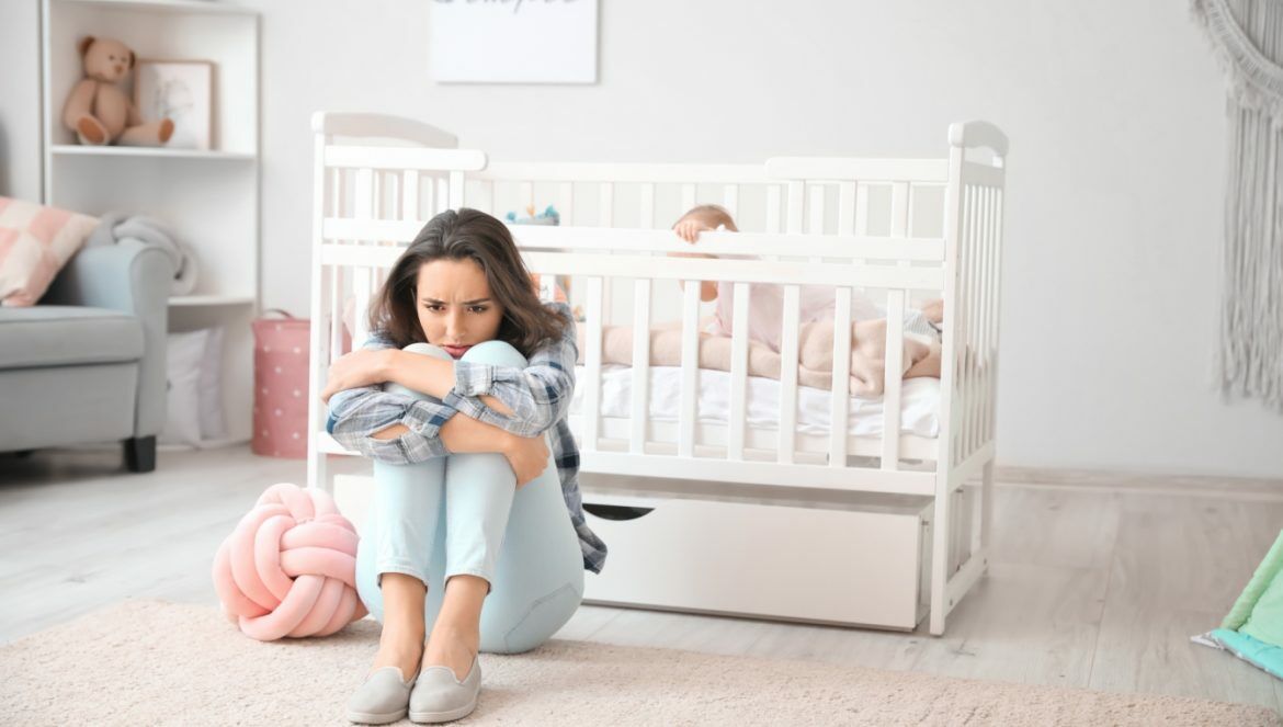 Depresja poporodowa - objawy i leczenie. Jak skutecznie wyleczyć depresję po porodzie? Smutna kobieta siedzi z podkulonymi nogami na środku dziecięcego pokoju, za nią stoi łóżeczko, w którym siedzi niemowlę.