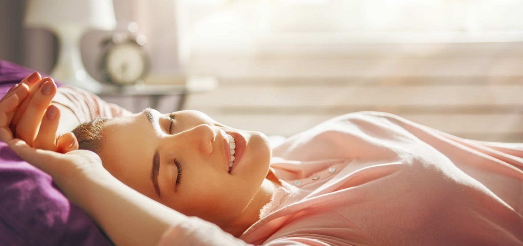 Sen na zdrowie - dlaczego nie warto zarywać nocy? 7 powodów, dla których warto się wysypiać. Kobieta w różowej bluzce przeciąga się w łóżku o poranku.