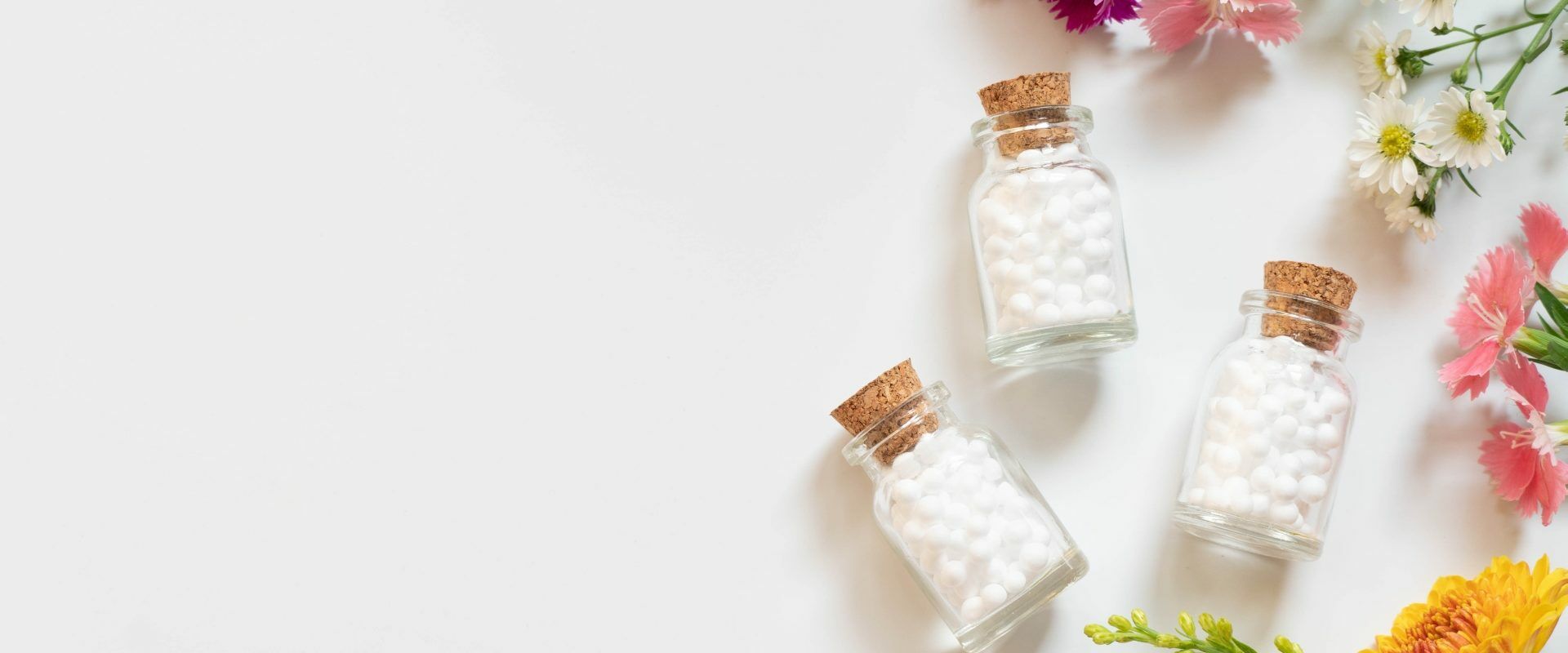 Homeopatia na niestrawność. Leki homeopatyczne w fiolkach, leżące na płasko na białym blacie. Obok leżą kwiaty.