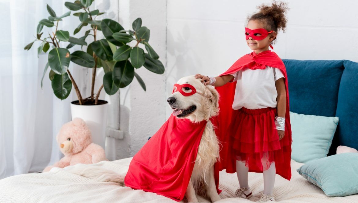 Dzieci i zwierzęta - przyjaźń, która procentuje.Jak pomóc dziecku zbudować dobrą relację ze zwierzakiem domowym? Mała dziewczynka i pies golden retriver ubrani w czerwone peleryny superbohaterów stoją na łóżku w sypialni rodziców.