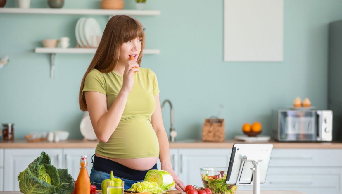 Dieta w ciąży - co powinna jeść przyszła mama? Kobieta w ciąży szykuje obiad w kuchni z ekologicznych produktów.