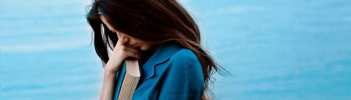 Jak poradzić sobie z depresją wg medycyny chińskiej? Smutna kobieta w niebieskiej sukience i długich czarnych włosach stoi nad brzegiem morza ze spuszczoną głową i przyciska do klatki piersiowej książkę.