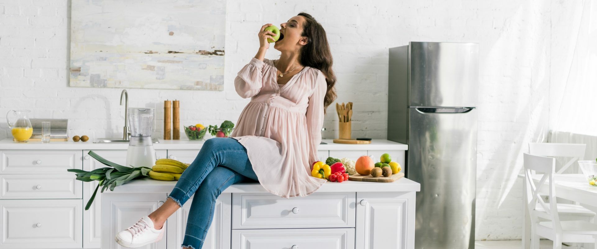Zgaga w ciąży - jak sobie z nią poradzić? Kobieta w ciąży siedzi na blacie w kuchni je jabłko.