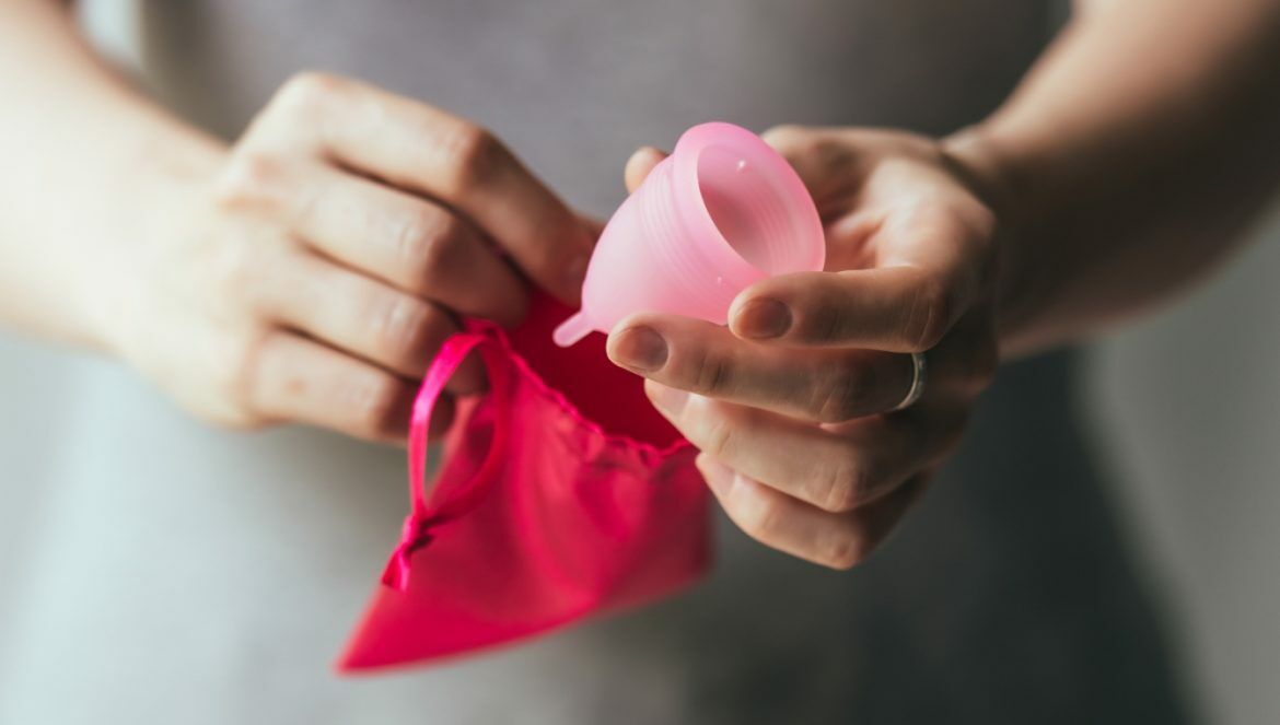 Kubeczek menstruacyjny i podpaski wielorazowe - na czym polega ekologiczna menstruacja? Kobieta w szarej sukience chowa różowy kubeczek do różowego woreczka.