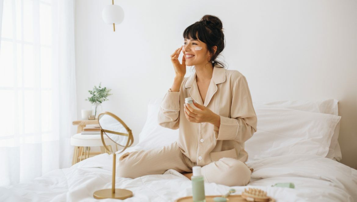 7 sposobów na naturalny demakijaż - jakie produkty roślinne pomagają dokładnie oczyścić twarz? Uśmiechnięta kobieta w beżowej piżamie siedzi na łóżku i pielęgnuje skórę twarzy.