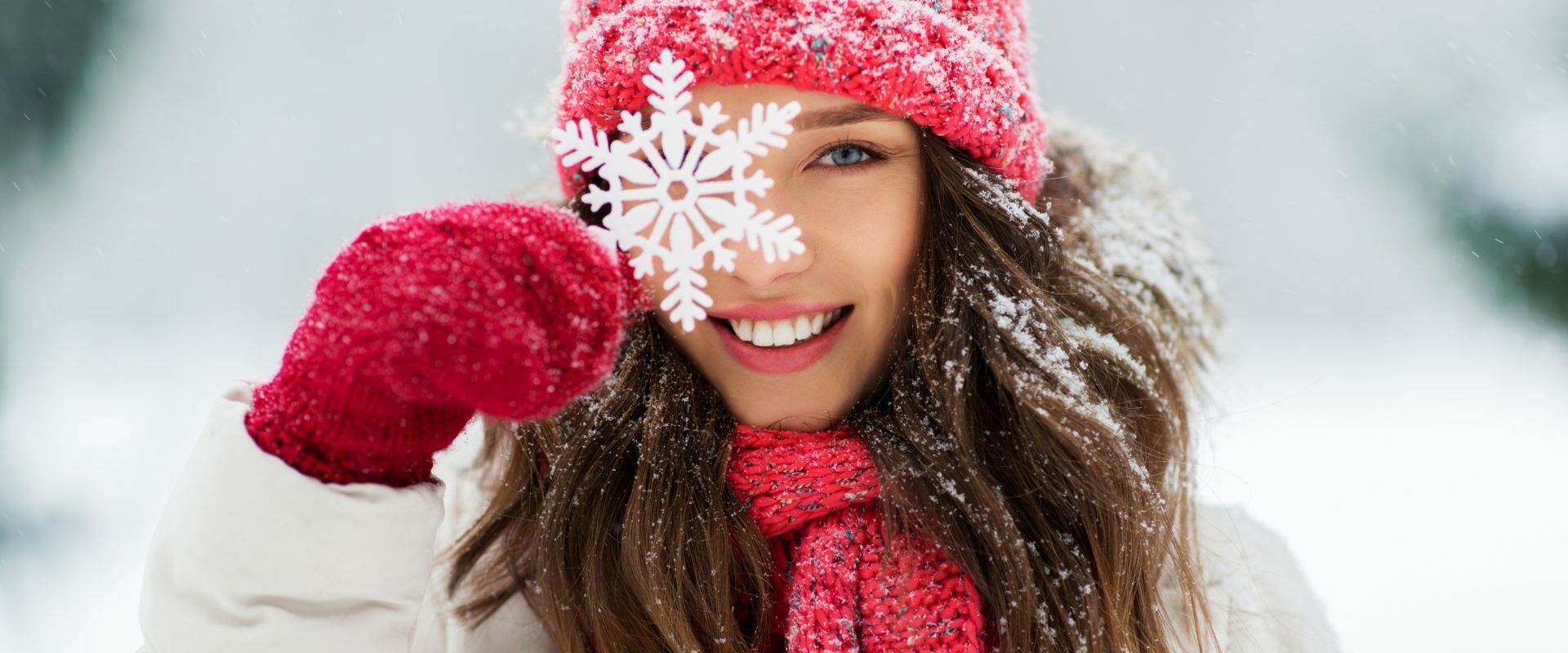 Jak wzmocnić odporność w 7 krokach? Kobieta w kurtce puchowej, czerwonej czapce i szaliku w zimowej scenerii uśmiecha się do aparatu, przykładając do oka ozdobny płatek śniegu.