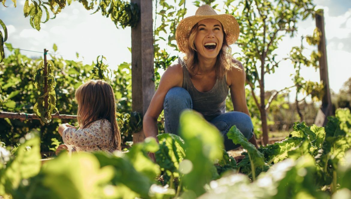 Ekologiczny ogród - jak go stworzyć od podstaw? Radosna kobieta w słomkowym kapeluszu z córką pracują w zielonym ogródku.