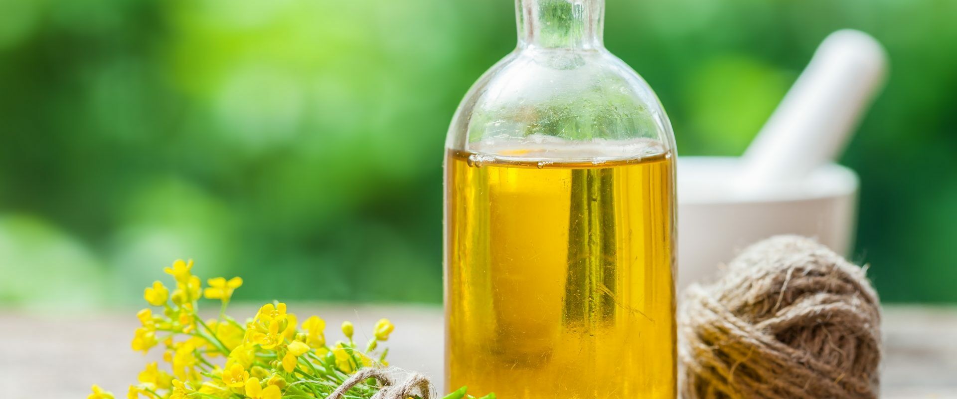 Olej rzepakowy - zdrowy czy szkodliwy? Olej rzepakowy w butelce, bukiet rzepaku i biały moździerz na drewnianym stole.
