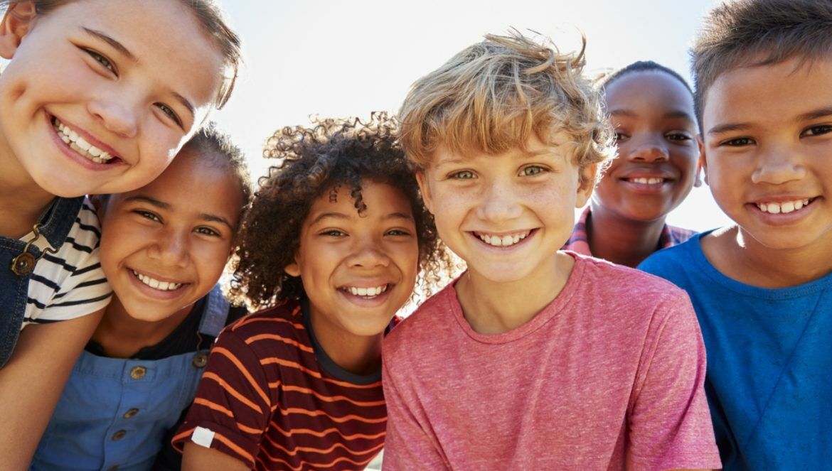 Jak nauczyć dziecko pozytywnego myślenia? Grupa uśmiechniętych dzieci o różnym pochodzeniu etnicznym patrzy w obiektyw aparatu.