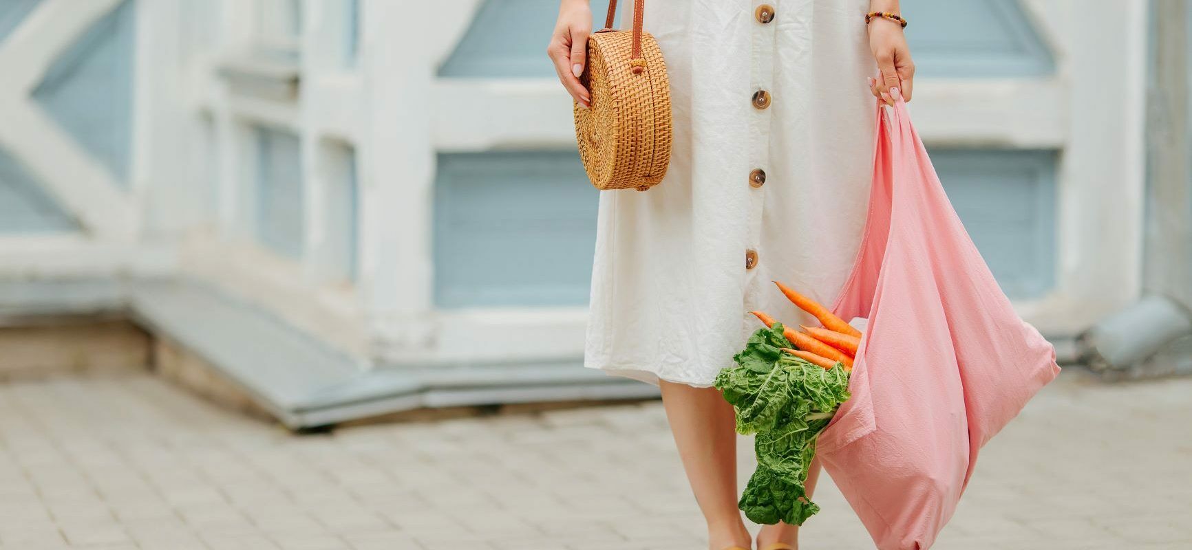 Jak być eko? Jak żyć ekologicznie? Kobieta w białej sukience i sandałach, z wiklinową torebką wiszącą na ramieniu stoi na tle białego budynku i trzyma w ręku różową materiałową torbę na zakupy z warzywami w środku.