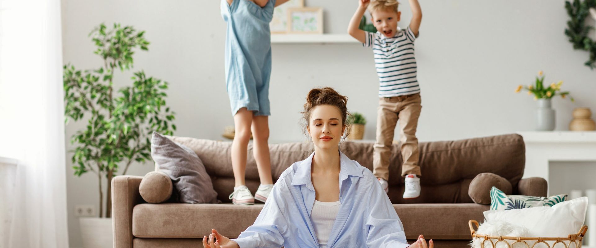 Work-life balance - jak odnaleźć równowagę? Młoda mama siedzi po turecku na podłodze w salonie i medytuje. Za nią na kanapie skaczą dzieci, obok stoi wiklinowy kosz z kocami i poduszką.
