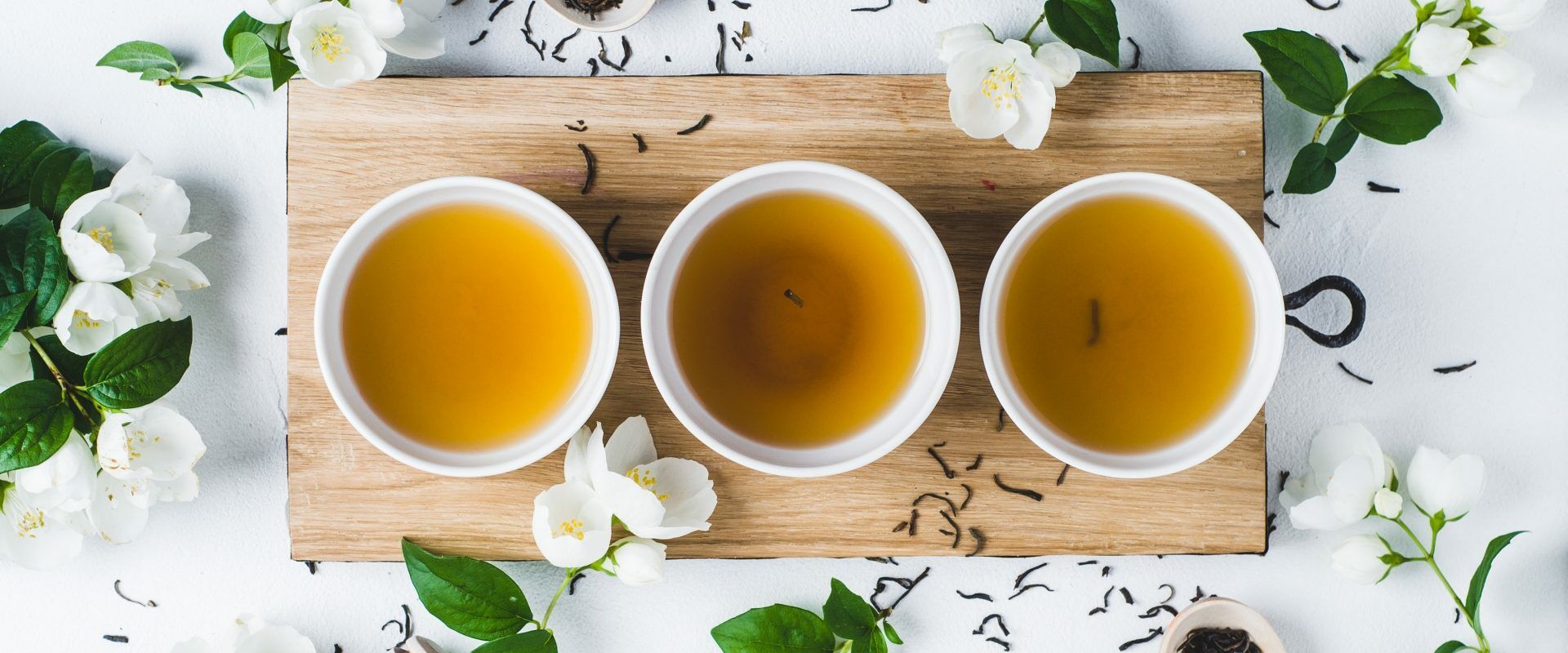 Zielona herbata - jakie domowe kosmetyki DIY można zrobić z zielonej herbaty? Zielona herbata w 3 filiżankach na drewnianej desce, wokół leżą listki herbaty i jaśmin.